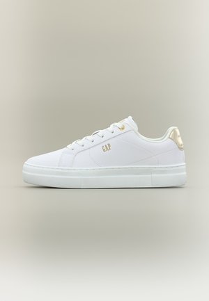 Hvit lavsko med hvite skolisser, gullfarget "GAP"-logo på siden og gullfarget detalj på hælfliken, vist mot en beige bakgrunn.