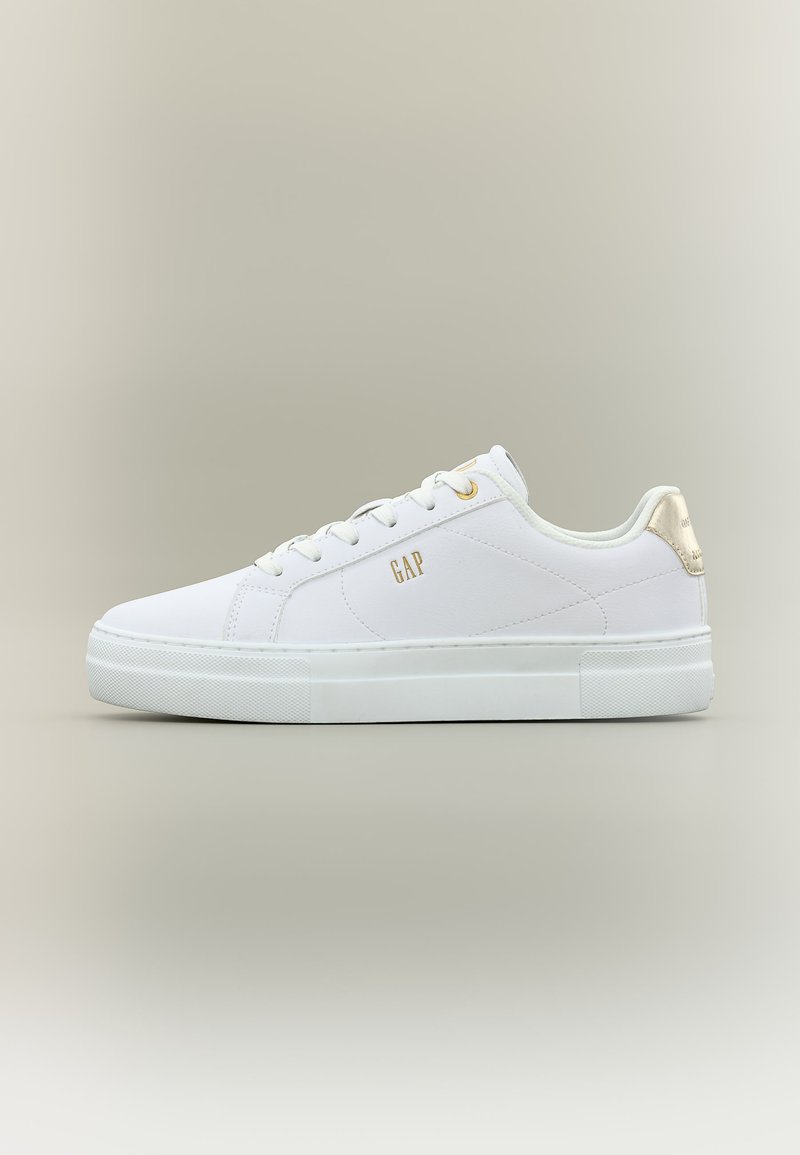 Baskets basses blanches avec lacets blancs, logo "GAP" doré sur le côté et accent doré sur la languette du talon, présentées sur un fond beige.
