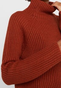 Knitted turtleneck sweater in a deep orange color, featuring ribbed texture, long sleeves, and a relaxed fit with visible seam details.
