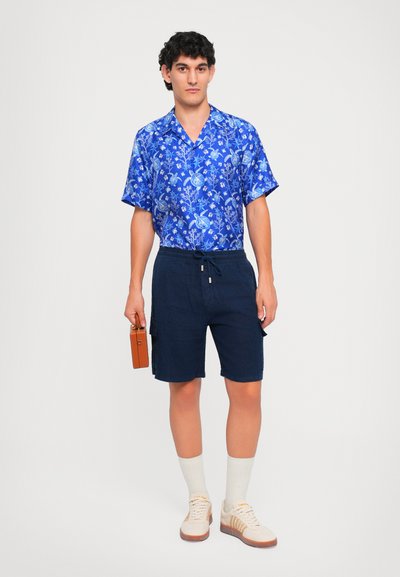 Lyhythihainen sininen kukkakuosisy shirt, tummansiniset sortsit, joissa on taskut, beigekengät ja valkoiset sukat. Kantaa pientä ruskeaa suorakaiteen muotoista laukkua.