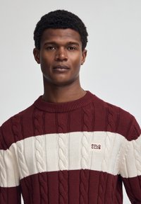 Kabelstickad tröja i vinröd och vit, med rund halsringning, horisontella ränder och en logotypärm för "POLO CLUB" på bröstet.
