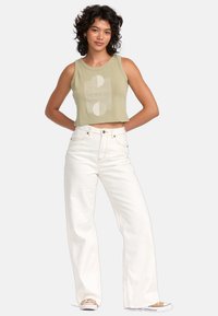 Afgeknotte olijfgroene tanktop met abstracte witte grafiek; gecombineerd met hoge witte flare jeans, allemaal gedragen met casual sneakers.