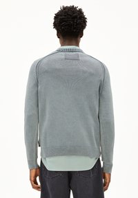 Lichtblauwe trui met geribde kraag en manchetten, gestructureerd breipatroon en een gelaagd effect over een mintgroen shirt, gezien vanuit de achterkant.