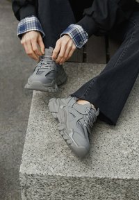 Szare buty sportowe mają teksturowany design z siateczkowymi panelami i wyraźnymi gumowymi podeszwami. Osoba nosząca je siedzi na betonowej powierzchni.