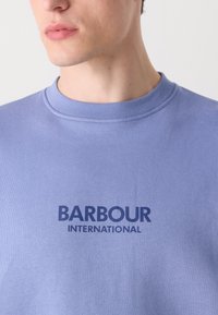 Világoskék pamut pulóver, kerek nyakkivágással és sötétkék "BARBOUR INTERNATIONAL" felirattal a mellkasán. Egyszerű, texturált anyag.