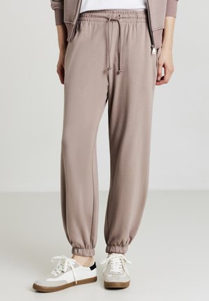 Pantaloni della tuta color taupe chiaro realizzati in tessuto morbido. Presentano un elastico in vita con cordoncino e polsini a costine. Abbinati a scarpe da ginnastica bianche e grigie.