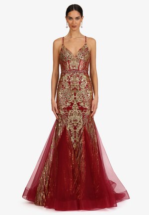 Frau trägt ein rotes Meerjungfrauenkleid mit goldener barocker Stickerei, dünnen Trägern und einem transparenten Tüllvolant am Saum, stehend vor weißem Hintergrund.