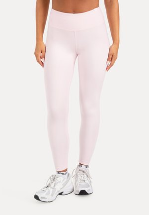 Vrouw draagt lichtroze high-waist leggings en witte sneakers met merksokken, staand tegen een effen achtergrond.