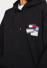 Sudadera negra con cordón, que presenta parches textiles coloridos con la inscripción "TOMMY JEANS" en varios colores y patrones en la parte delantera.