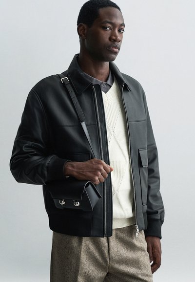 Giubbotto bomber in pelle nera con polsini a coste, chiusura a zip e tasche laterali. Abbinato a un maglione strutturato color crema e pantaloni a quadri marroni.