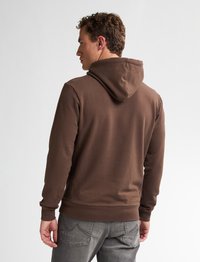 Felpa in cotone marrone con cappuccio regolabile, polsini e orlo a costine. La vista posteriore mostra una superficie liscia e una vestibilità casual, abbinata a jeans grigi.
