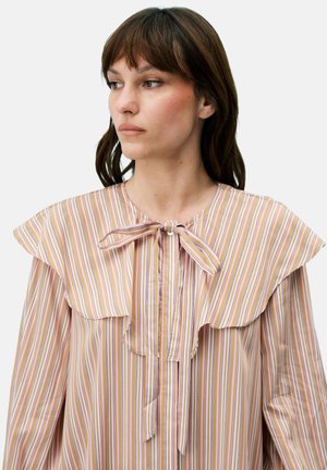 Femme aux cheveux bruns portant un chemisier rayé verticalement beige, rose et blanc avec un grand col à volants et un nœud noué au niveau de l'encolure.