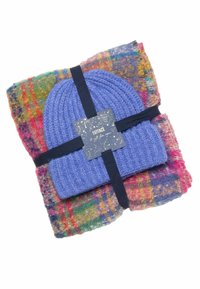 Blue knit beanie atop a colorful, patterned plaid scarf, secured with a dark ribbon and a decorative tag. Soft materials with varied textures.