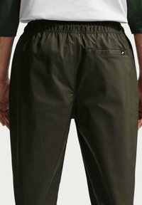 Vue de dos d'une personne portant un pantalon Nike vert foncé avec une ceinture élastique et un petit logo sur la poche arrière droite.