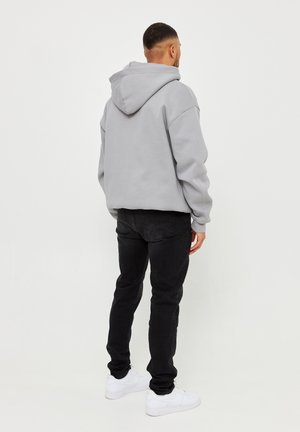 Sudadera gris con capucha y hombros caídos, combinada con jeans negros y zapatillas blancas. La tela parece suave y tiene un ajuste relajado.