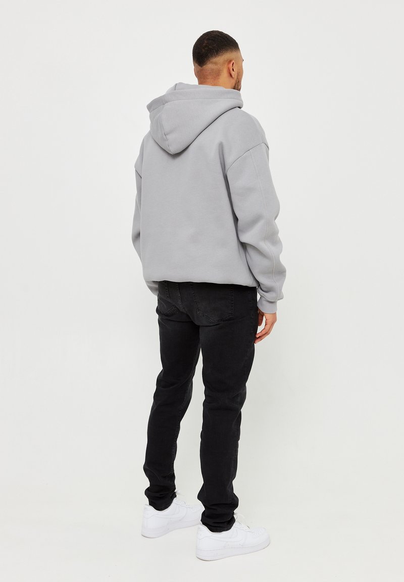 Felpa grigia con cappuccio e spalle cadute, abbinata a jeans neri e sneakers bianche. Il tessuto appare morbido con una vestibilità comoda.