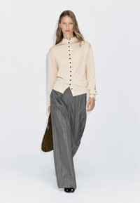 Gilet beige avec des boutons noirs, doté d'un col montant, associé à un pantalon large à rayures grises. Le mannequin tient un sac marron.