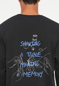 Cilvēks ar melnu kreklu, uz kura ir zils teksts "Daloties pie galda, veidojot atmiņas" un baltas līnijas zīmējumi ar rokām, kas spēlē kārtis un glāzēm uz muguras.