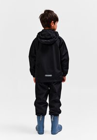 Czarna wodoodporna kurtka z kapturem, w zestawieniu z czarnymi spodniami i niebieskimi gumowcami. Tkanina o fakturze, luźny fason oraz widoczne logo na plecach.