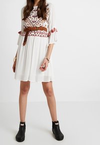 Vestido blanco con detalles bordados en naranja y marrón, con mangas acampanadas y un cinturón. Botas negras hasta el tobillo completan el look.