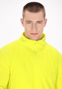 Felpa in pile giallo fluorescente con colletto alto e chiusura a mezza zip. Texture morbida e polsini senza cuciture, ideale per visibilità e calore.