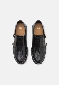 Chaussures en cuir verni noir à semelle plate, avec deux brides à boucle et des détails décoratifs à la brogue sur le bout et les côtés.