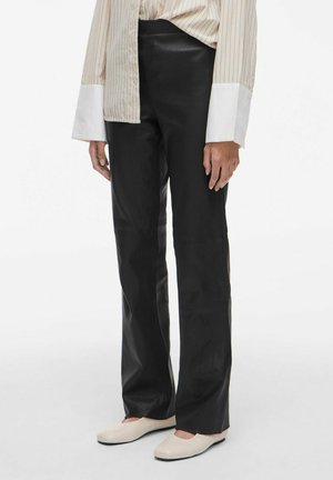 Femme portant un pantalon droit en cuir noir, une chemise rayée beige avec de larges poignets blancs et des chaussures plates de couleur crème.