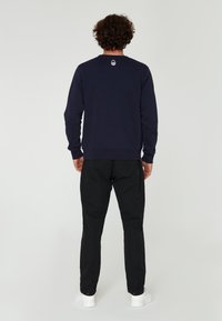 Sudadera azul marino con cuello redondo, puños de canalé y logo en la parte trasera, combinada con pantalones negros y zapatillas blancas. Diseño minimalista.