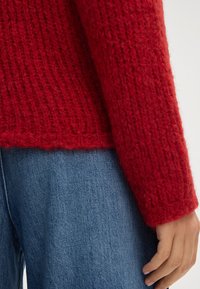 Maglione rosso a maglia con un motivo testurizzato, caratterizzato da una vestibilità rilassata e polsini a coste, abbinato a jeans blu. Dettaglio di filato morbido e peloso visibile.