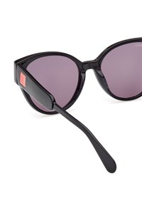 Gafas de sol negras en forma de ojo de gato con lentes morados, que presentan un acabado brillante y un acento de logo rojo en el costado. Las patillas negras delgadas se extienden hacia afuera.