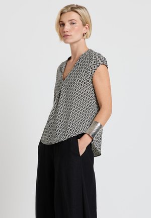 Donna con capelli biondi corti che indossa una blusa senza maniche a motivo geometrico bianco e nero e pantaloni neri, mano in tasca, bracciale a polsino d'argento.