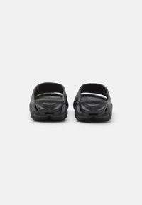Par de sandalias negras tipo slip-on con suelas texturizadas y talones abiertos, mostradas desde la parte posterior sobre un fondo blanco.