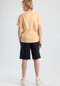 Peach cotton t-shirt and black shorts. T-shirt has short sleeves and a round neck. Shorts have a pocket and reach mid-thigh. White sneakers worn.