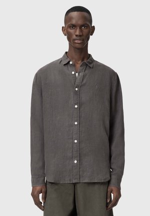 Mężczyzna ubrany w długorękawową ciemnoszarą koszulę na guziki i oliwkowozielone spodnie, stojący na tle jasnoszarego tła.