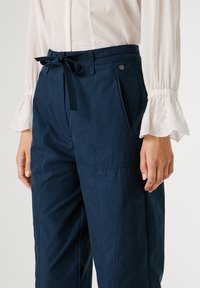 Παντελόνια σκούρου μπλε χρώματος με υφή, εξοπλισμένα με κορδέλα στη μέση, πλαϊνές τσέπες και ορατές ραφές, φορεμένα με λευκό πουκάμισο.