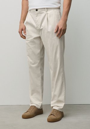 Mann trägt beige plissierte Hose und braune Wildleder-Schnürschuhe und steht auf einem glatten Boden vor einer schlichten Wand.