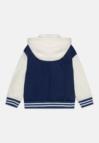 Veste marine et blanche avec capuche, présentant des manches contrastantes, des poignets élastiques et un ourlet côtelé avec des rayures. Texture lisse, matériau léger.