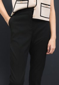 Fekete, testhez simuló nadrág sima textúrával és rejtett oldalzsebekkel, bézs színű ujjatlan felsővel párosítva, amely fekete hangsúlyokkal és gombokkal rendelkezik.