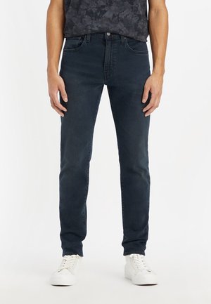 Homme portant un jean slim bleu foncé et des baskets blanches, debout devant un fond blanc uni.