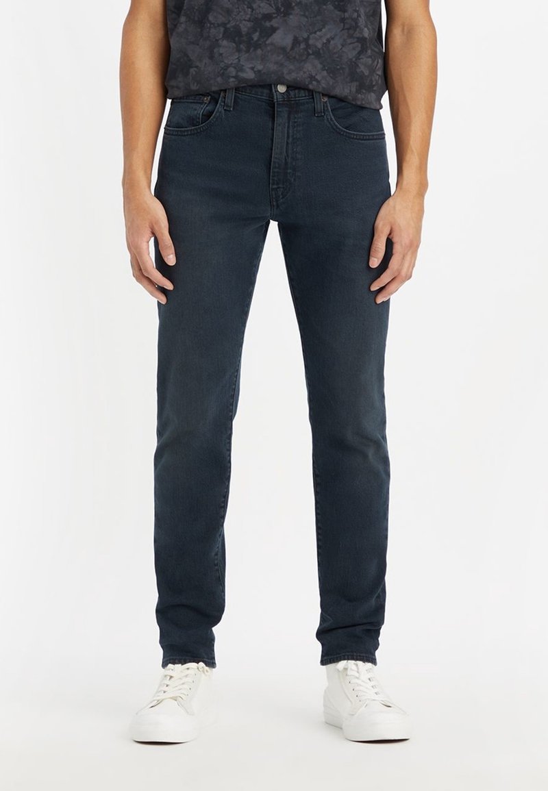 Homme portant un jean slim bleu foncé et des baskets blanches, debout devant un fond blanc uni.