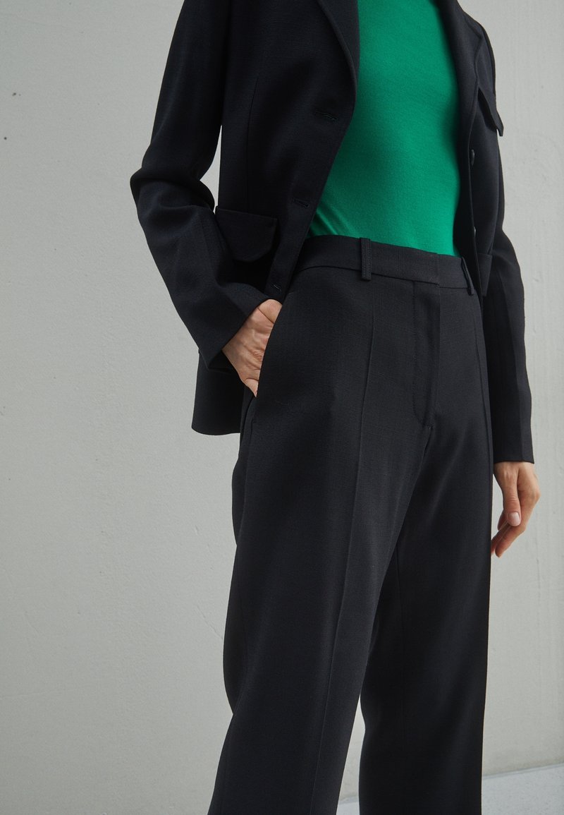 Svart skräddarsydd kavaj i kombination med slanka svarta byxor och en figurnära grön topp. Slätt tyg, strukturerad design med fickor på kavajen.