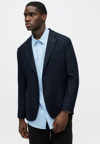 Blazer navy con un corte entallado, cierre de un solo botón y dos bolsillos, sobre una camisa de botones a rayas azul claro. Acabado en tela suave.