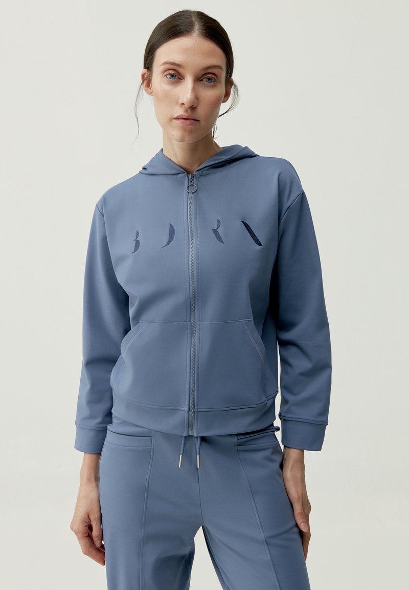 Blaue Zip-Hoodie aus glattem Stoff, mit einer Brusttasche und dezenten Logodetails. Begleitet von passenden Jogginghosen.