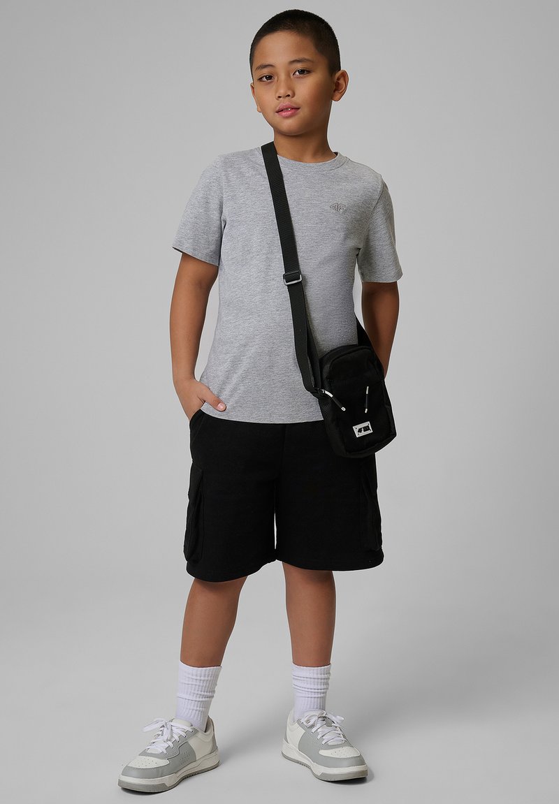 Garçon portant un t-shirt gris, un short noir, des baskets blanches et un sac bandoulière noir, debout devant un fond uni clair.