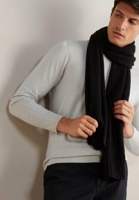 Sort tørklæde lavet af struktureret stof, hængt om halsen. Sammen med en lys grå sweater og mørke bukser, der viser et enkelt design.