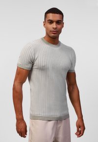 Hellgrauer, gestrickter Kurzarmpullover mit strukturiertem Wellenmuster und grünem Saum am Kragen und den Ärmeln, kombiniert mit hellen Hosen.