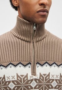 Camisola castanha de malha com gola alta, com fecho de correr e um padrão geométrico de floco de neve em creme e azul-marinho na parte inferior.