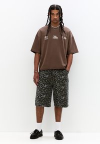 Oversized brown cotton t-shirt with white logos, paired with knee-length gray shorts featuring a dark leopard print. Black shoes and white socks.