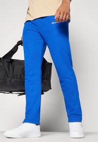 Blå joggebukser med en komfortabel passform, elastisk midjebånd og en liten hvit logo. Kombinert med hvite sneakers og en svart duffelbag.