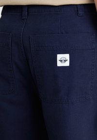 Navy blue cotton pants feature dual back pockets with a white label displaying "Dockers" and a logo. Fabric is smooth with a subtle sheen.